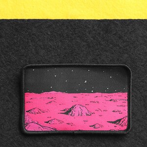 May include: A pink and black embroidered patch depicting a landscape of a planet with a starry night sky. The patch is surrounded by a black border with a white stitch.