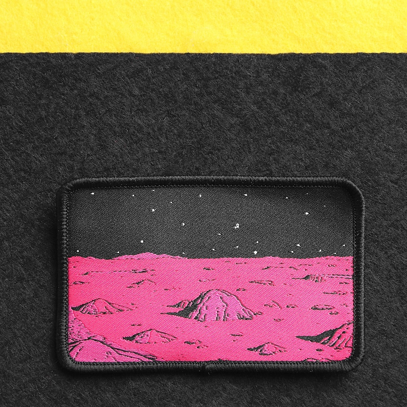 May include: A pink and black embroidered patch depicting a landscape of a planet with a starry night sky. The patch is surrounded by a black border with a white stitch.