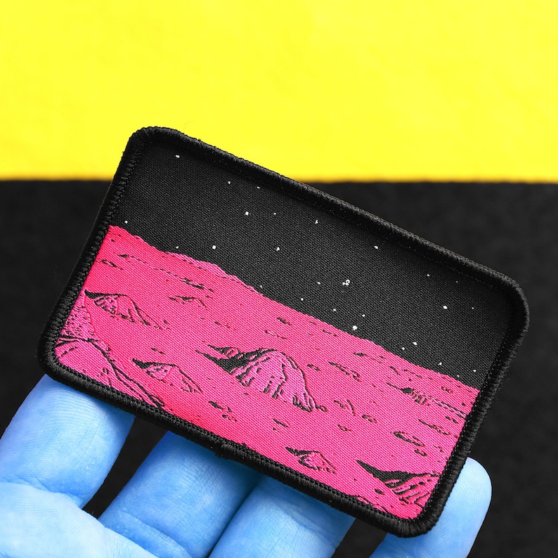 May include: A black and pink embroidered patch depicting a landscape of a planet with craters and a starry sky. The patch has a black border and is made of fabric.