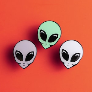 May include: Three alien face pins, one green, one white and one light purple. The aliens have black eyes and a black smile.