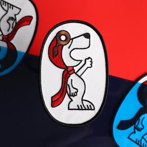 May include: White embroidered patch featuring a cartoon dog wearing a red aviator hat and scarf. The dog is standing on its hind legs with its mouth open and one paw raised.