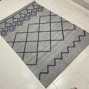 May include: A rectangular gray rug with a geometric design. The rug features a pattern of dark blue diamonds, squares, and zigzags. The rug is made of a woven material and is suitable for a living room or bedroom.