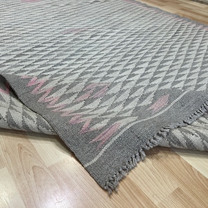May include: A grey and pink patterned rug with a fringe border. The rug has a geometric design with a diamond pattern.