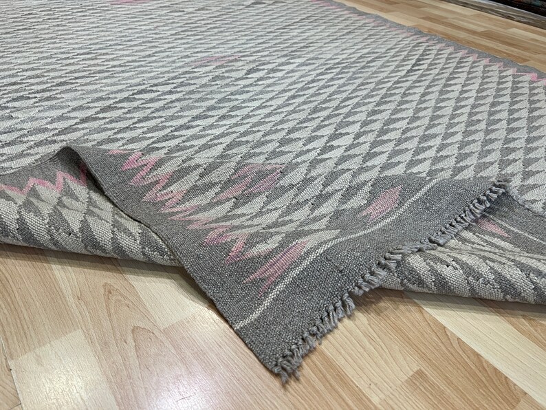 May include: A grey and pink patterned rug with a fringe border. The rug has a geometric design with a diamond pattern.