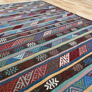 Puede incluir: Una alfombra rayada colorida con un patrón geométrico. La alfombra tiene un fondo marrón con rayas azules, verdes, rojas y blancas.