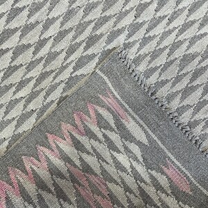 May include: Close-up of a grey and white woven rug with a pink and white geometric pattern. The rug has a fringed edge.