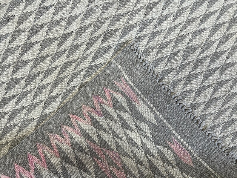 May include: Close-up of a grey and white woven rug with a pink and white geometric pattern. The rug has a fringed edge.