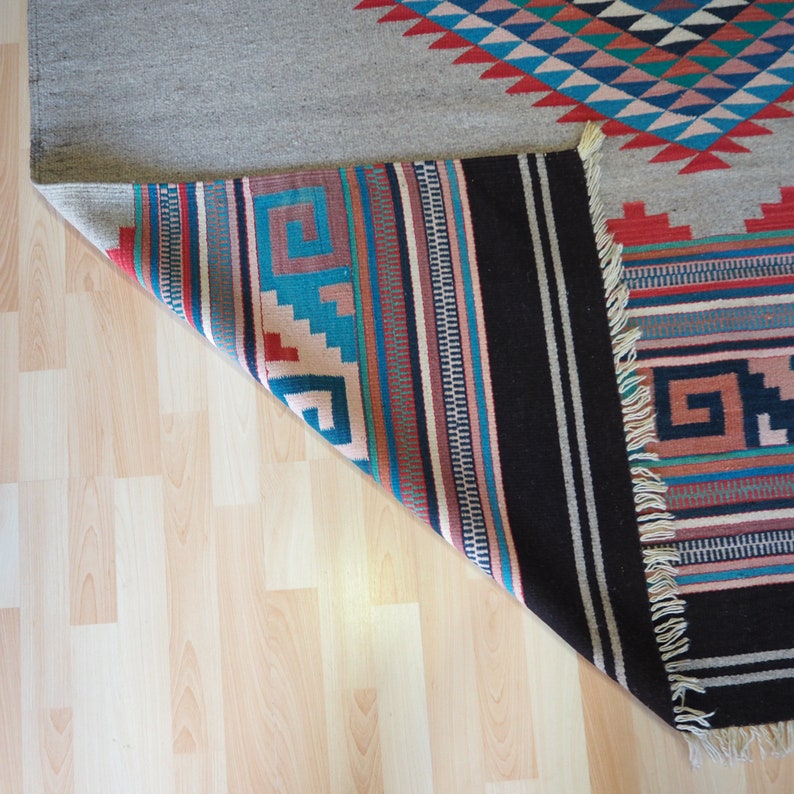 May include: A close-up of the underside of a colorful woven rug with fringe. The rug has a geometric pattern in shades of red, blue, and white. The rug is folded over, revealing a black and white striped pattern.