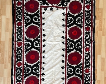 May include: A white fabric textile with a black and red embroidered border. The border features a repeating pattern of stylized flowers and circles.