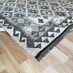 May include: A geometric patterned rug in shades of gray, white, and black. The rug features a repeating square design with triangular and diamond shapes. The edge of the rug is visible, showing a fringe detail. The rug is on a light wood floor.