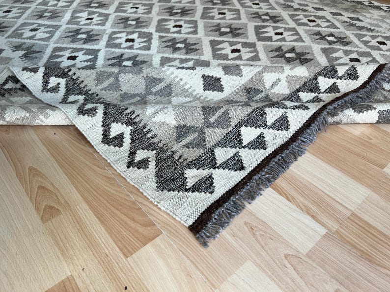 May include: A geometric patterned rug in shades of gray, white, and black. The rug features a repeating square design with triangular and diamond shapes. The edge of the rug is visible, showing a fringe detail. The rug is on a light wood floor.