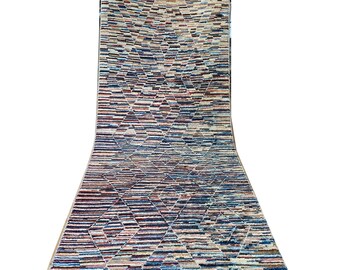 Op de afbeelding: Een lang, rechthoekig tapijt met een geometrisch ruitpatroon. Het tapijt heeft horizontale strepen in tinten blauw, beige, rood en bruin. Het tapijt is opgerold, wat de lengte en het ontwerp laat zien.