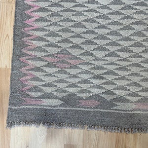 May include: A grey and pink woven rug with a geometric pattern. The rug has a fringed border.