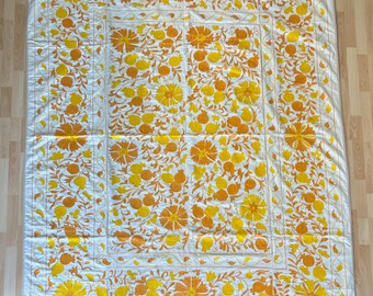 May include: A white quilt with a repeating floral pattern in shades of yellow and orange. The quilt features large stylized flowers and fruit, with a border of similar design. The overall aesthetic is bright and cheerful, suitable for a bedroom or living room.