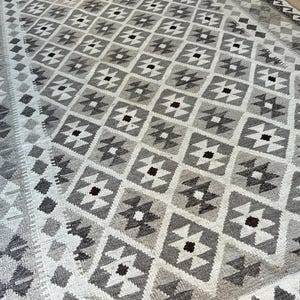 May include: A large, rectangular rug with a geometric pattern in shades of gray and white. The design features repeating diamond shapes with smaller, contrasting accents. The rug has a border with a detailed pattern.