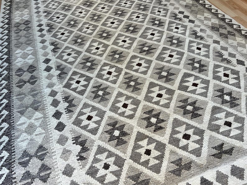 May include: A large, rectangular rug with a geometric pattern in shades of gray and white. The design features repeating diamond shapes with smaller, contrasting accents. The rug has a border with a detailed pattern.
