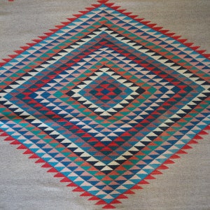 May include: A Navajo rug with a geometric pattern of red, blue, green, and white triangles. The rug is woven with a diamond shape in the center.