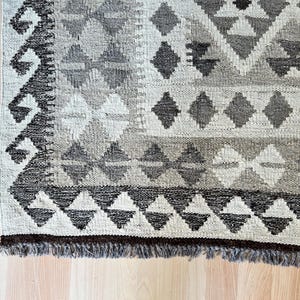 May include: Close-up of a hand-woven rug with a geometric pattern. The rug features diamond and triangle shapes in shades of gray, beige, and brown. The rug has a fringed edge and is placed on a light-colored wooden floor.