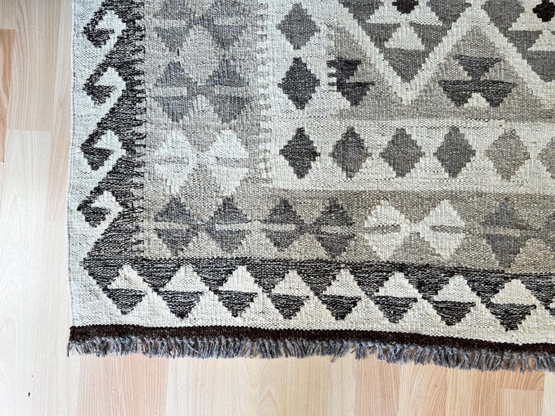 May include: Close-up of a hand-woven rug with a geometric pattern. The rug features diamond and triangle shapes in shades of gray, beige, and brown. The rug has a fringed edge and is placed on a light-colored wooden floor.