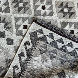 May include: A close-up of a gray and white patterned rug with a geometric design. The rug features a repeating pattern of squares, stars, and diamonds in shades of gray, white, and brown. The rug has a fringed edge.