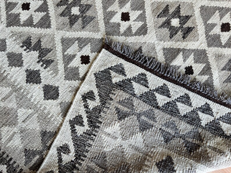 May include: A close-up of a gray and white patterned rug with a geometric design. The rug features a repeating pattern of squares, stars, and diamonds in shades of gray, white, and brown. The rug has a fringed edge.