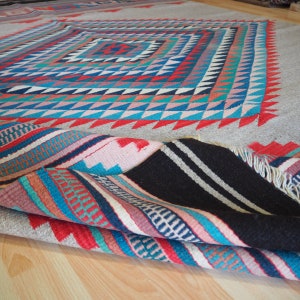 May include: A colorful handwoven rug with a geometric pattern in shades of red, blue, pink, and white. The rug is folded in half, revealing the intricate details of the woven design.