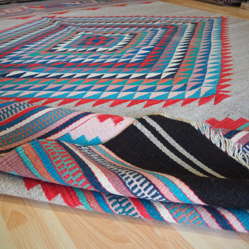 May include: A colorful handwoven rug with a geometric pattern in shades of red, blue, pink, and white. The rug is folded in half, revealing the intricate details of the woven design.