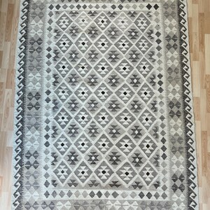 May include: Rectangular rug with a geometric diamond pattern in shades of gray and cream. The rug has a border with a zigzag design and fringe on the short ends. The rug is made of woven material.