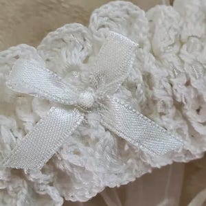 May include: Close-up of a white, crocheted garter with a small satin bow. The garter features a delicate, openwork design and is likely intended for a wedding or special occasion. The bow adds a touch of elegance.