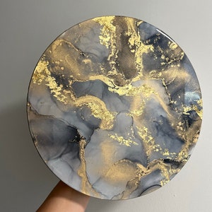 May include: A round, gray and gold resin art piece with a marbled design. The piece has a white background with gray and black swirls and gold flecks.