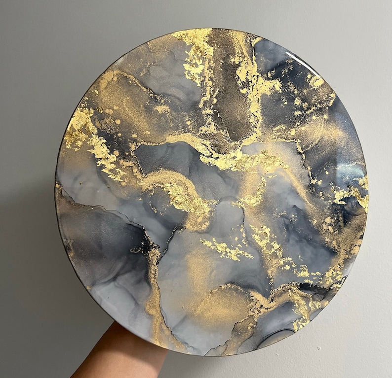 May include: A round, gray and gold resin art piece with a marbled design. The piece has a white background with gray and black swirls and gold flecks.