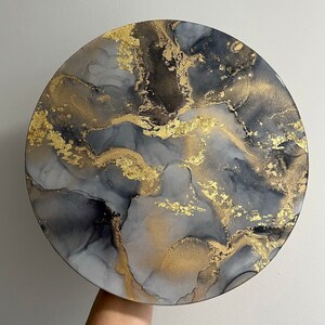 May include: A round, gray and black abstract painting with gold accents. The painting has a marbled effect and is reminiscent of a galaxy.