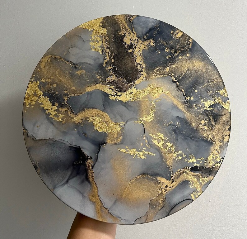 May include: A round, gray and black abstract painting with gold accents. The painting has a marbled effect and is reminiscent of a galaxy.