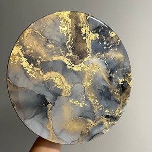 May include: Round resin art piece with a black, gray, and gold abstract design. The design resembles swirling marble.