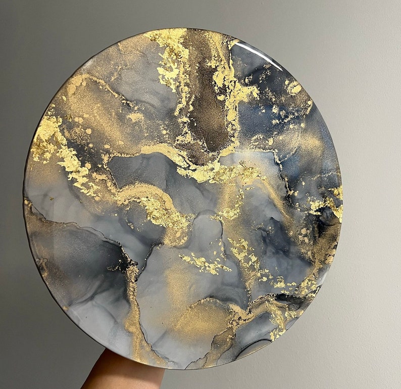 May include: Round resin art piece with a black, gray, and gold abstract design. The design resembles swirling marble.