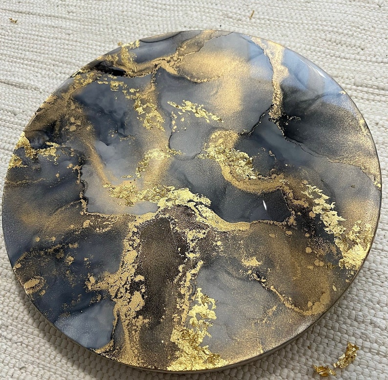 May include: A round, black and gold resin tray with a marbled design. The tray has a smooth, glossy finish and is decorated with gold leaf.