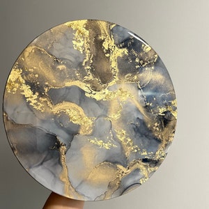 May include: A round, gray and gold resin art piece with a marbled design. The piece has a white background with gray and gold swirls and flecks.