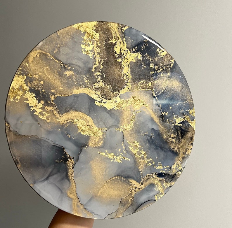 May include: A round, gray and gold resin art piece with a marbled design. The piece has a white background with gray and gold swirls and flecks.