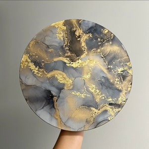 May include: A round, gray and gold abstract painting with swirling patterns. The painting has a white background with black and gold swirls.