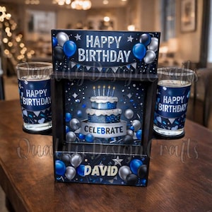 May include: A birthday-themed display box with two matching glasses. The box features the text "Happy Birthday" and "Celebrate", along with a cake illustration and the name "David". Blue and silver balloons decorate the box and glasses.