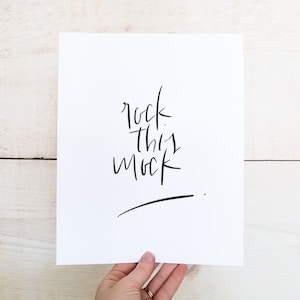 May include: A white card with black calligraphy that reads "rock this mock" on a white wood background.