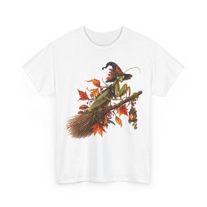 May include: A white t-shirt featuring a graphic illustration of a green praying mantis wearing a black pointed hat, perched on a broomstick decorated with autumn leaves, small mushrooms, berries, and a hanging lantern with a warm glow.