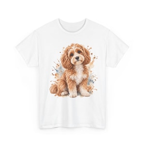 Op de afbeelding: Een wit T-shirt met korte mouwen en een centrale grafische illustratie van een pluizige, goudbruine en witte puppy die zit. De hond is omringd door artistieke aquarelachtige verfspatten in tinten bruin, beige en blauw.