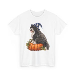 May include: A white t-shirt featuring a graphic illustration of a black and white dog wearing a blue wizard hat with yellow stars and a crescent moon, sitting on top of a large orange pumpkin with green leaves and curling vines at the base.