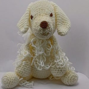Peut inclure: Un chien au crochet blanc cassé fait à la main, avec des yeux et un nez bruns. Le chien a des oreilles tombantes et un corps hirsute et texturé. Le chien est assis droit, mettant en valeur ses pattes crochetées et son design général.