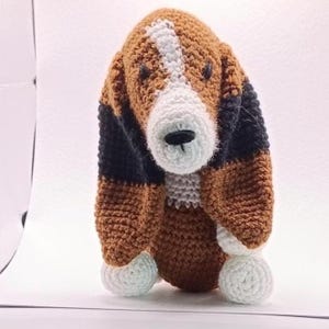 May include: A crochet stuffed animal shaped like a dog with long ears. The toy features a tri-colour pattern of brown, black, and white yarn. It has a white stripe running down the centre of the face, a black stitched nose, and white paws.