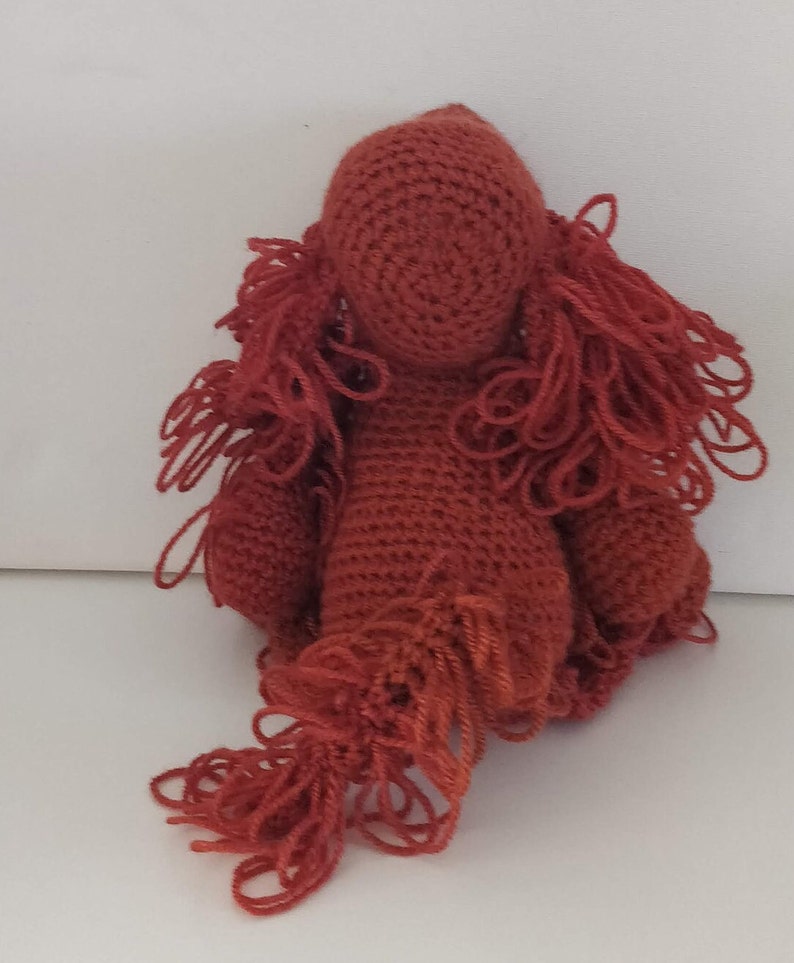 May include: A crocheted red plush toy shaped like a dog with long, shaggy hair. The toy has a simple face with no eyes or mouth.