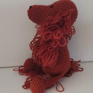 May include: A crocheted red lion plush toy with a fluffy mane. The lion is sitting with its head turned to the side.
