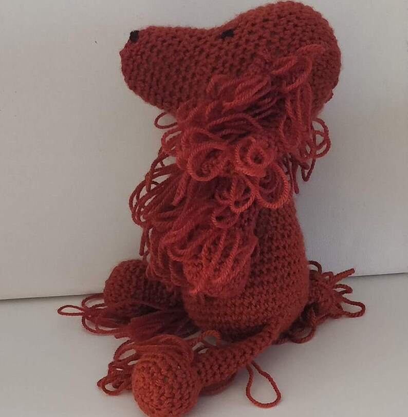 May include: A crocheted red lion plush toy with a fluffy mane. The lion is sitting with its head turned to the side.