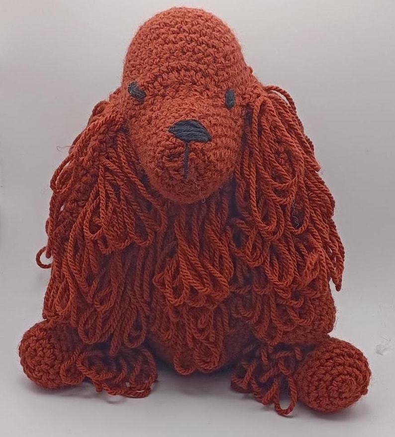 May include: A crocheted red dog toy with a long, shaggy mane. The dog is sitting with its legs tucked under its body.
